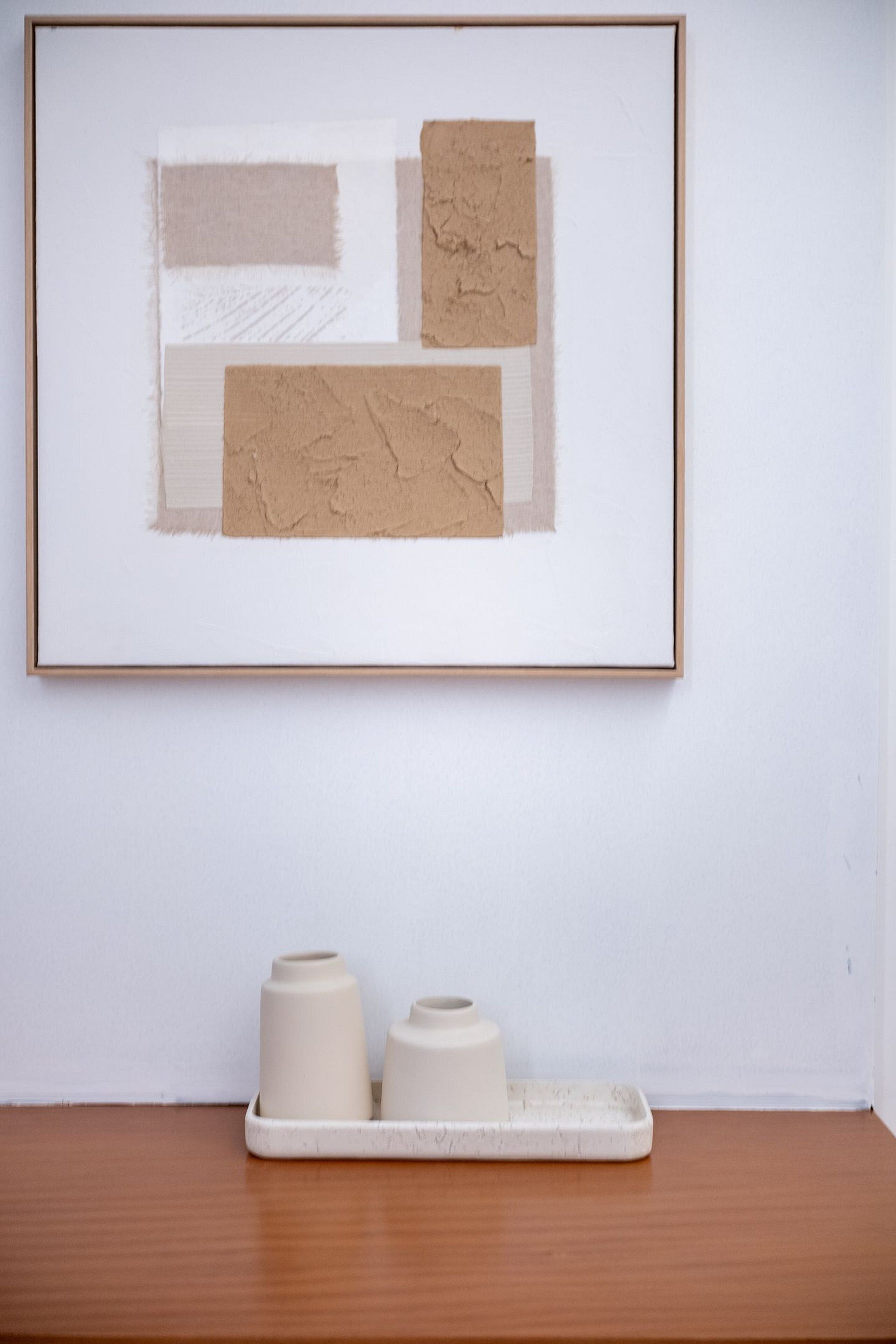 A framed artwork with abstract shapes and textures on a wall. Below it, a tray holds two cylindrical objects.
