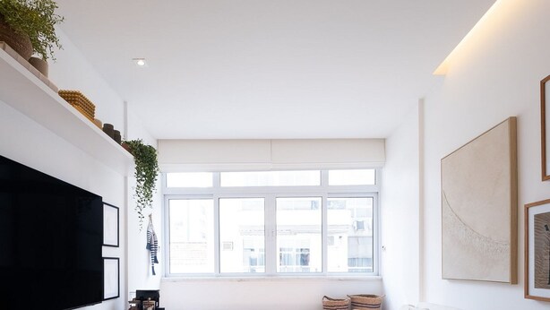 A modern living room with a large flat-screen TV, a sofa, a window with a view, and a wall-mounted shelf with plants. - Impeccable apartment in a great location in Leblon (Rio de Janeiro)
