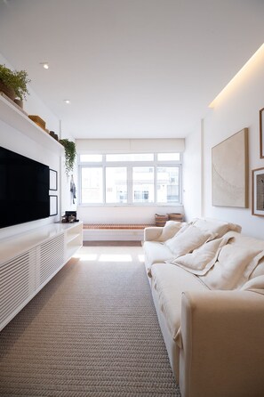 A modern living room with a large flat-screen TV, a sofa, a window with a view, and a wall-mounted shelf with plants. - Impeccable apartment in a great location in Leblon (Rio de Janeiro)