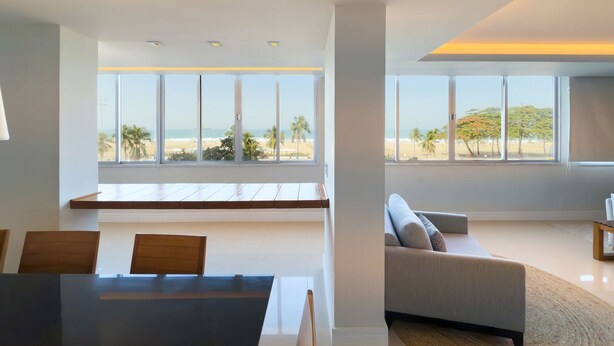 A modern living room with a large window offering a view of the beach. There is a dining area with a table and chairs. - Copacabana | Av. Atlântica, Oceanfront, 4 Bedrooms (Rio de Janeiro)