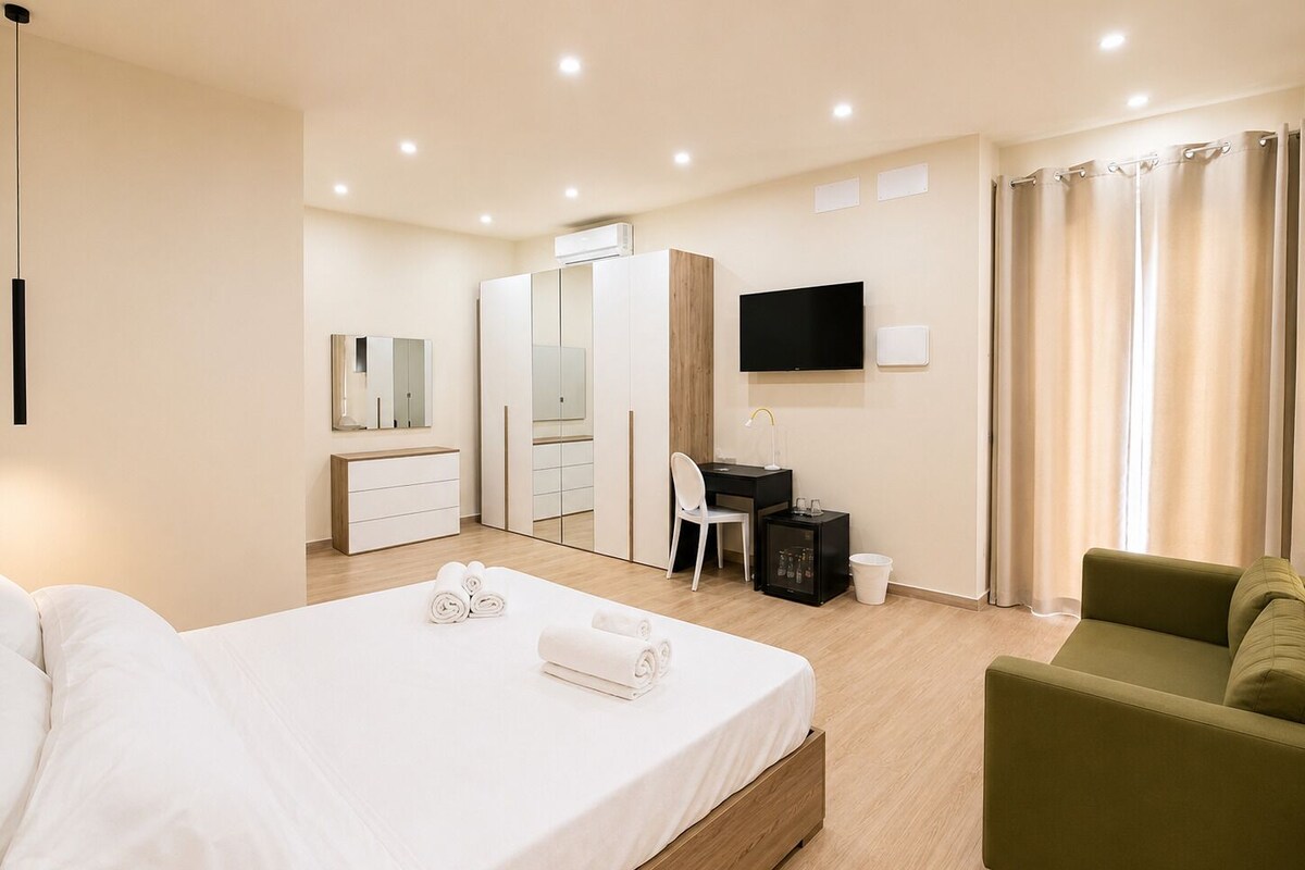 Quarto de hotel com cama, escrivaninha, cadeira, televisão e sofá verde.