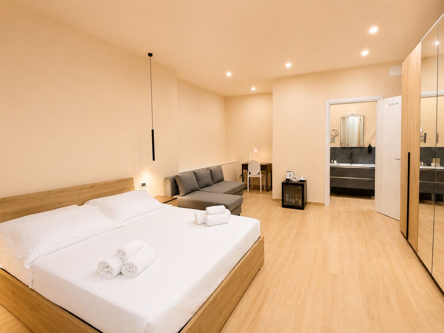 A modern hotel room with a large bed, a sofa, a desk, and a bathroom with a mirror and sink.