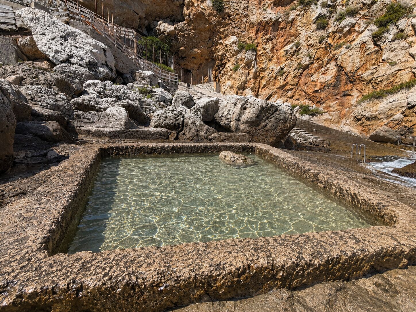 Une source d’eau chaude naturelle, entourée d’un paysage rocheux et accessible par un escalier en bois.