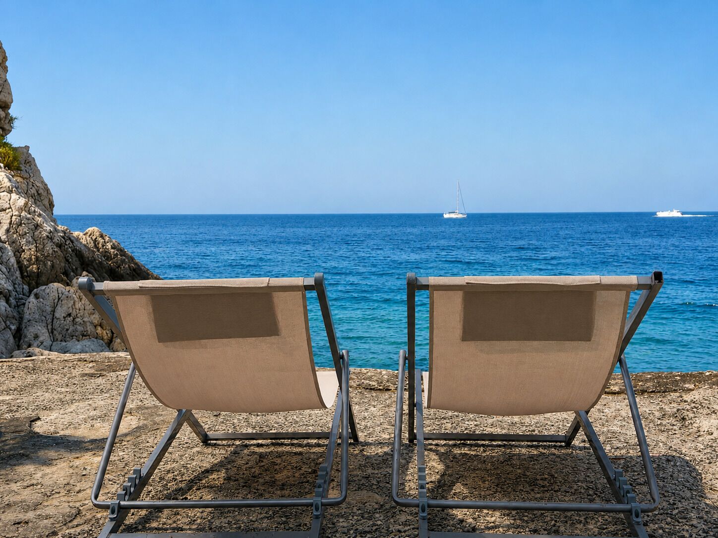 Two beach chairs facing the sea with rocky terrain and a clear sky.