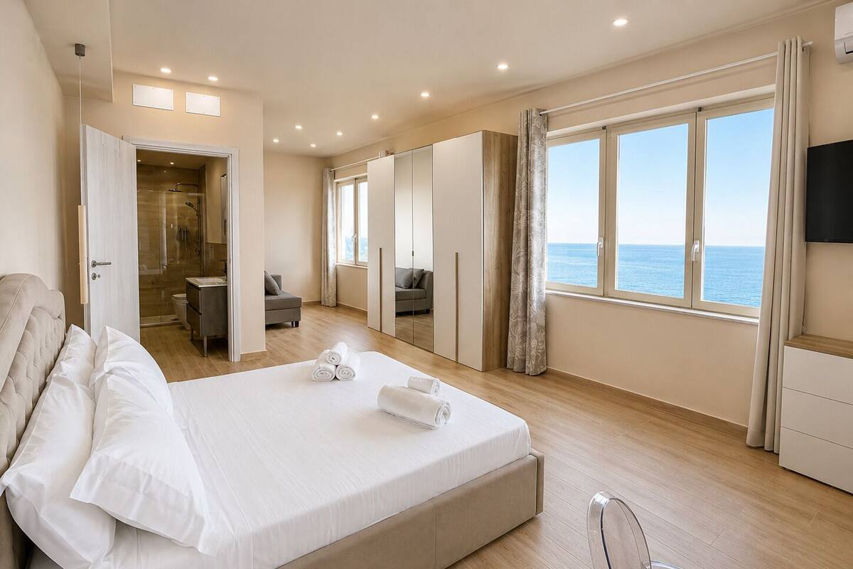 Une chambre à coucher comprenant un lit, un bureau, une chaise, une armoire et une grande fenêtre donnant sur la mer.