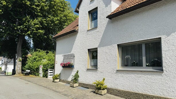 Un bâtiment blanc avec un toit recouvert de tuiles, une fenêtre ornée d’un motif en forme de cœur et des plantes en pot disposées le long du mur.
