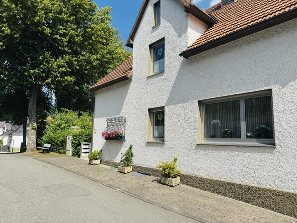 A white building with a tiled roof, a window with a heart-shaped decoration, and potted plants along the side.