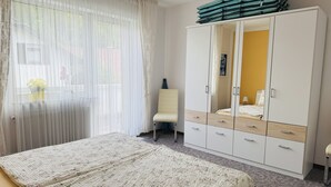 Une chambre à coucher comprenant un lit, une armoire, une chaise et une fenêtre avec des rideaux légers.