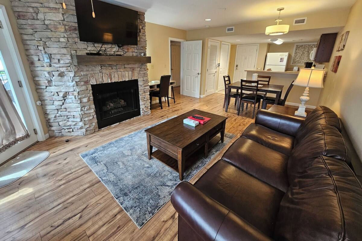 A living room with a stone fireplace, leather sofa, wooden coffee table, and a dining area with chairs and a table.