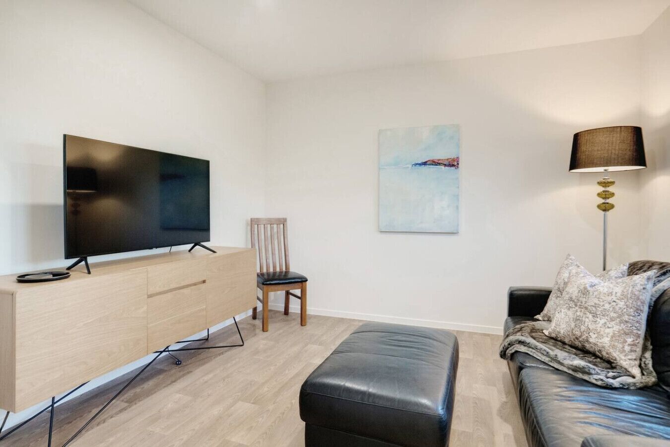 Een moderne woonkamer met een flatscreen televisie op een houten dressoir, een zwart leren poef, een zwarte bank met gekleurde kussens, een houten stoel en een vloerlamp.