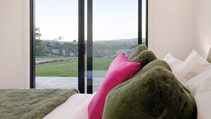 Un dormitorio con un ventanal grande, una cama con almohadas verdes y rosas, y vistas a un paisaje verde.