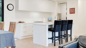 Una cocina moderna con armarios blancos, una isla central y taburetes de bar. Hay un reloj en la pared y un cartel con un mensaje divertido.