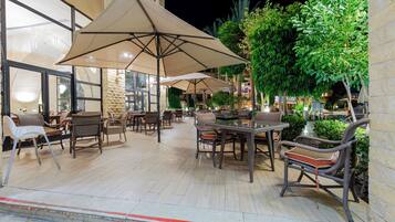 An outdoor seating area with tables and chairs, large umbrellas, and greenery.