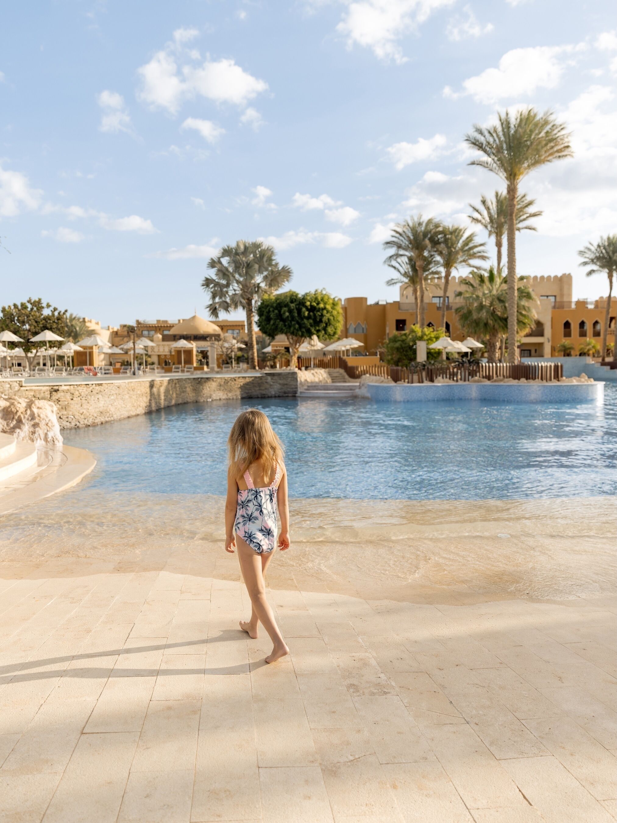 A pool area with a person walking, palm trees, and traditional architecture.