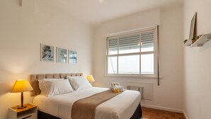 A neatly arranged bedroom with a bed, bedside tables, a window with blinds, and wall-mounted artwork. - Ae302 Zona1 in Rio de Janeiro (Rio de Janeiro)