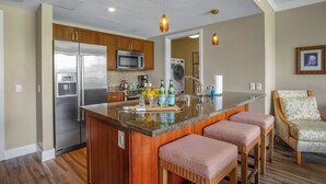 A modern kitchen with a central island, stainless steel appliances, wooden cabinets, and a seating area with wicker chairs. - Honua Kai | Ocean View 5 BR, Sleeps 16 | Car Incl. w/ 6+ Nights | HKH ML-1610 by KBM (Lahaina)