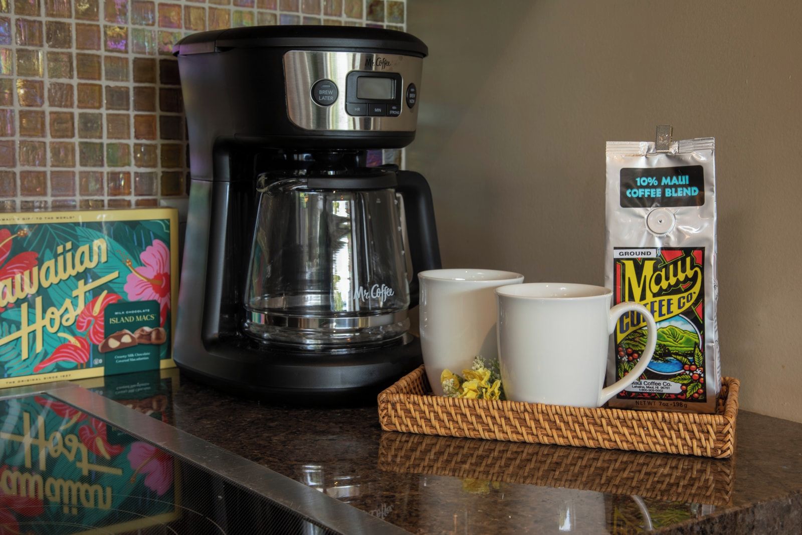 A coffee maker, two white mugs, a box of Hawaiian Host Island Macchiato, and a bag of Maui Coffee Blend on a kitchen counter.