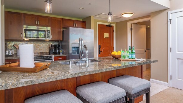 A kitchen with wooden cabinets, stainless steel appliances, a granite countertop, bar stools, and hanging pendant lights. - Honua Kai | Ocean View 5 BR, Sleeps 16 | Car Incl. w/ 6+ Nights | HKH ML-1616 by KBM (Lahaina)