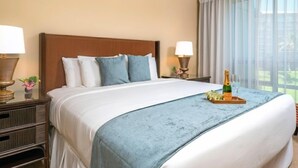 A hotel room with a large bed, two bedside tables with lamps, a window with curtains, and a bottle of champagne on the bed.