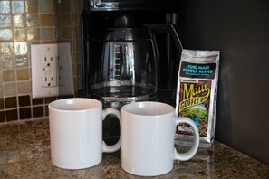 Dos tazas blancas sobre una encimera de granito, una cafetera negra y un paquete de café Maui Blend.