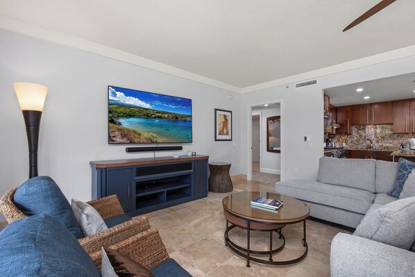 A living room with a flat-screen TV, a sofa, a coffee table, and a kitchen in the background.