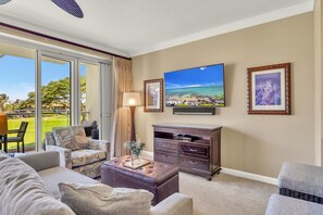 A living room with a large TV, a sofa, a coffee table, and a view of a golf course through the sliding glass door.