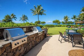 An outdoor grill area with stone walls, a view of the ocean, and a grassy lawn with palm trees.