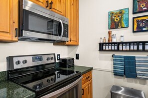 A kitchen with wooden cabinets, a stainless steel microwave, an electric stove, and a toaster. There are wall-mounted spice jars and a framed bear painting.