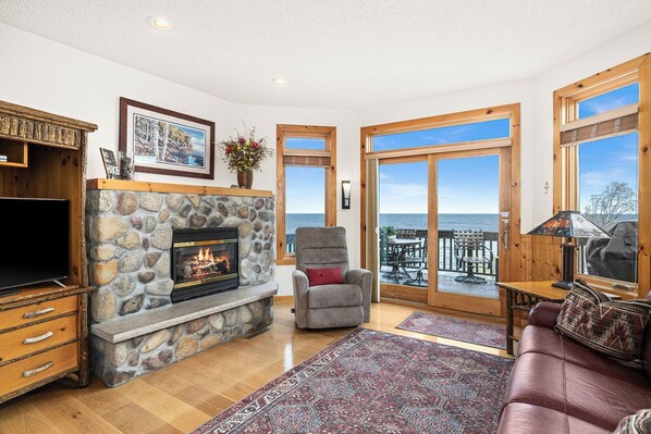 A living room with a stone fireplace, a television, a comfortable seating area, and a view of the ocean through large sliding doors.