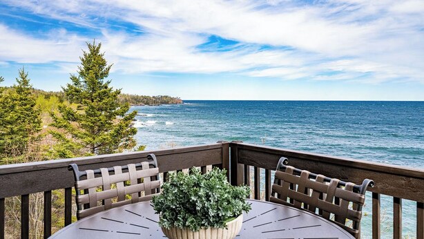 A wooden deck with a round table and chairs, overlooking a body of water and a forested coastline.