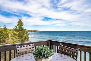 A wooden deck with a round table and chairs, overlooking a body of water and a forested coastline.