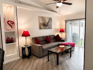 A modern living room with a grey sofa, red cushions, a glass coffee table, and a ceiling fan.