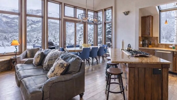 Un vaste salon doté d’une grande fenêtre donnant sur un paysage montagneux enneigé, un coin repas en bois et une cuisine avec un îlot central.