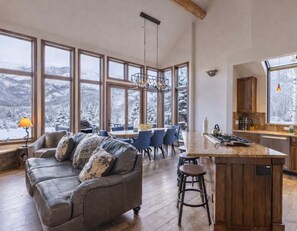 A spacious living room with a large window overlooking a snowy mountain landscape, a wooden dining area, and a kitchen with a central island. - Mountain Village Chalet | Walk to Gondola | Views (Mountain Village)
