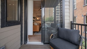 A balcony with a sofa and a chair, a sliding glass door leading to an interior space with a sofa and a lamp, and a view of a brick building. - PFR Suites - Highland, 3BR 2BA Shadyside Condo, 2 Decks and Off-Street Parking (Pittsburgh)