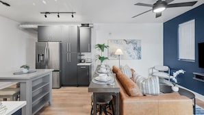 A modern kitchen with stainless steel appliances, a dining area with a wooden table and bar stools, and a living area with a sofa and a ceiling fan. - PFR Suites - Highland, 3BR 2BA Shadyside Condo, 2 Decks and Off-Street Parking (Pittsburgh)