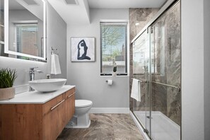A modern bathroom with a large mirror, a white sink, a wooden vanity, a toilet, and a glass shower enclosure. - Pristine Double King 3BR suite | A+ Location| ADA| (Pittsburgh)