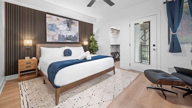 A modern bedroom with a bed, bedside tables, a ceiling fan, a large abstract painting, and a view of the outdoors through a glass door. - Immaculate Double King Suite | 4 beds | 2 Decks (Pittsburgh)