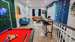 A pool table with balls, a blue sofa, an orange sofa, a dining table with chairs, and a staircase with string lights. - Royaleux / 4 King Beds / Sleep 16 / Pool Table (Philadelphia)