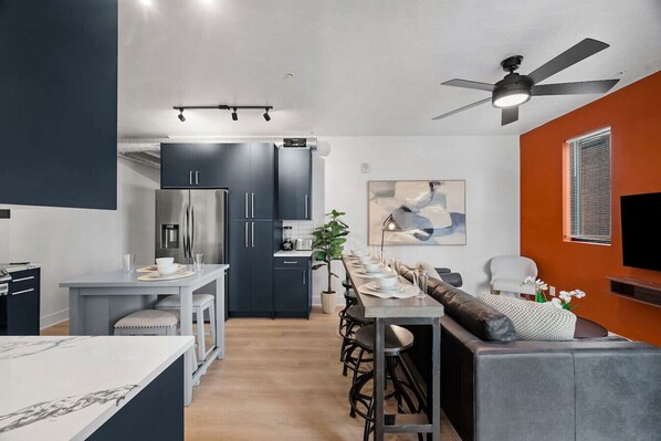 A modern kitchen with dark cabinets, a central island, and a dining area with a long table and bar stools.