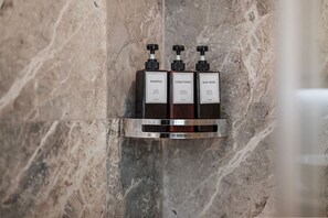 A bathroom with a marble wall and a shelf holding three bottles of shampoo, conditioner, and body wash.