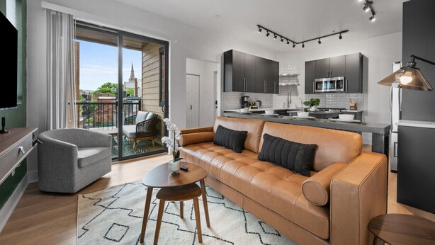 A modern living room with a leather sofa, a coffee table, and a TV. There's a balcony with a view of the city. - Stunning 3BR Penthouse Suite| 2 decks| A+ Location (Pittsburgh)