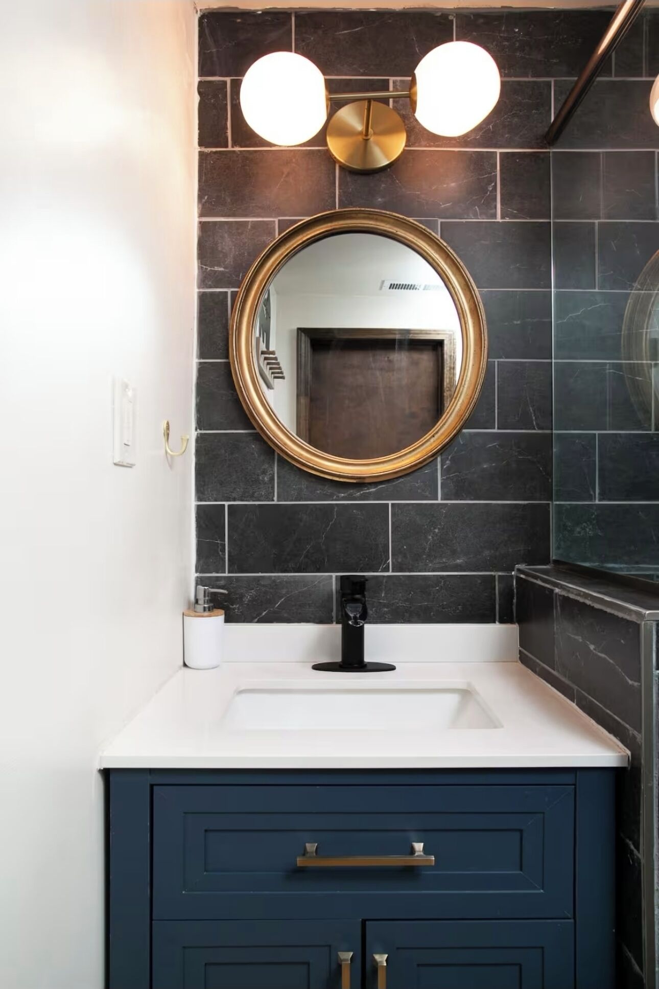 A bathroom with a dark blue vanity, a white countertop, a gold-framed mirror, and black marble tiles.