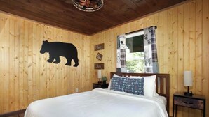 Ein Zimmer mit Holzwänden, einem Bett, einer Bärensilhouette an der Wand und einem Fenster mit einem gemusterten Vorhang.