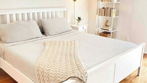 Ein ordentlich bezogenes Bett mit gestreifter Bettwäsche und einer Strickdecke, ein Nachttisch mit Lampe, ein Bücherregal und ein gerahmtes Bild an der Wand.