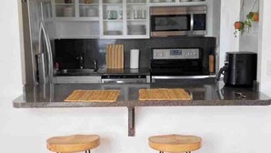 Eine Küche mit Edelstahl-Kühlschrank, Mikrowelle und Backofen, zwei hölzernen Barhocker und einer weißen Spritzschutzwand.