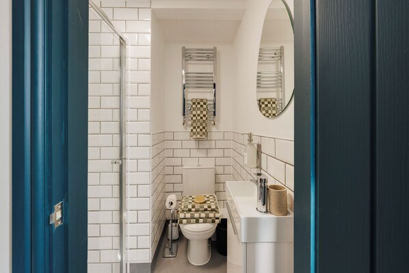 Un baño con paredes de azulejos blancos, un inodoro con tapa a cuadros, un perchero, un espejo y un lavabo con dispensador de jabón.