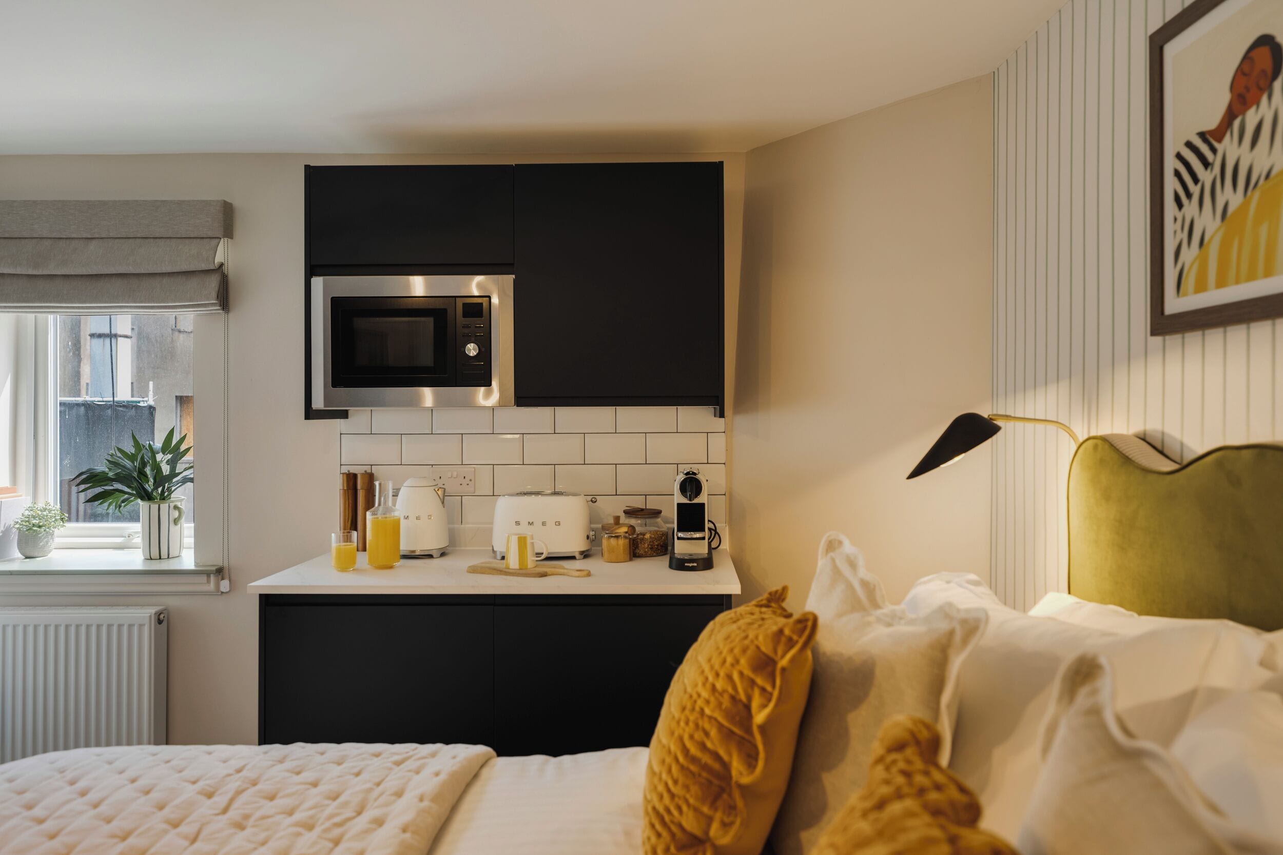 A modern kitchen with a microwave, toaster, and coffee maker. There is a bed with a yellow pillow and a green headboard.