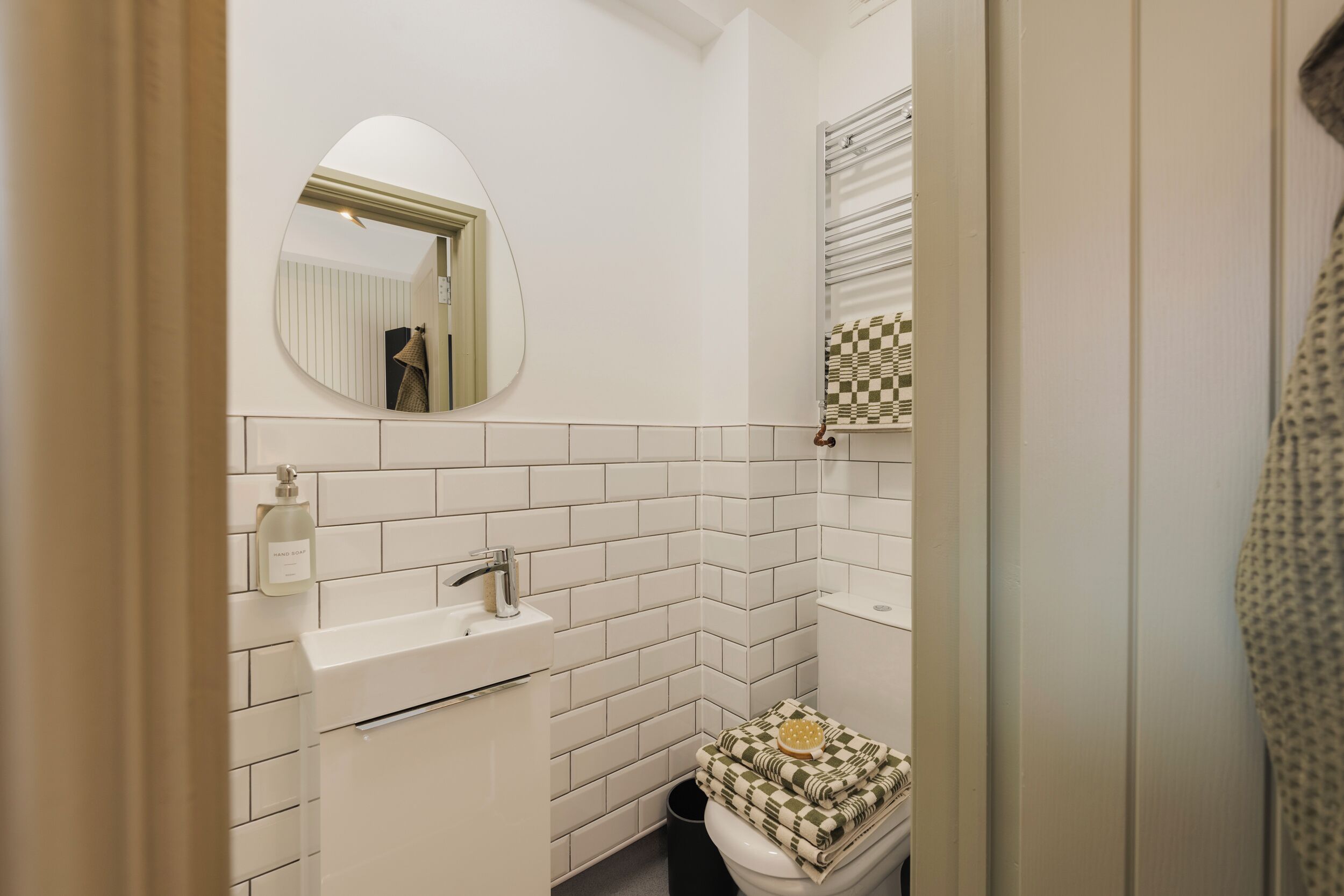 Baño con paredes de azulejos blancos, un espejo redondo, un lavamanos y un inodoro con tapa a cuadros.