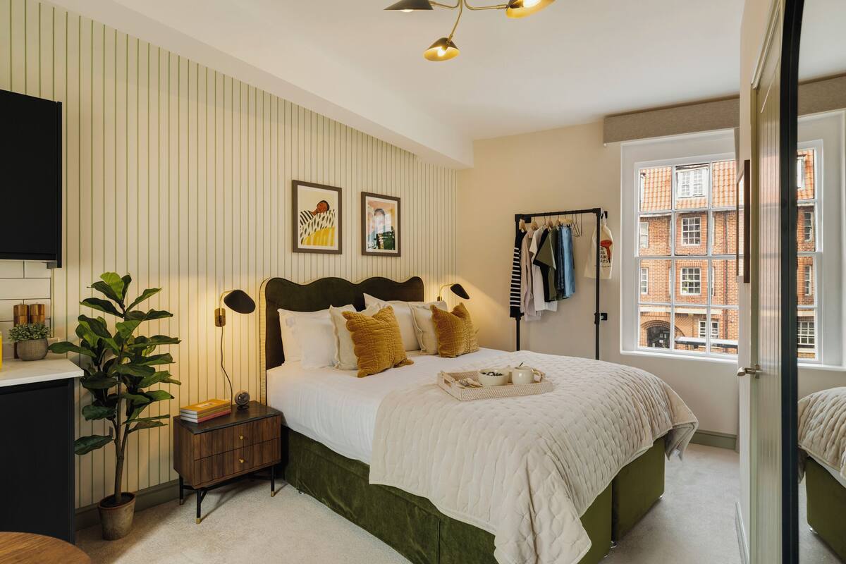 Un dormitorio con cama, mesita de noche, lámpara y vista a un edificio de ladrillo a través de una ventana.
